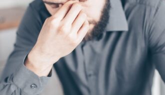 Bearded man looking stressed and tired, sitting at a desk with paperwork.