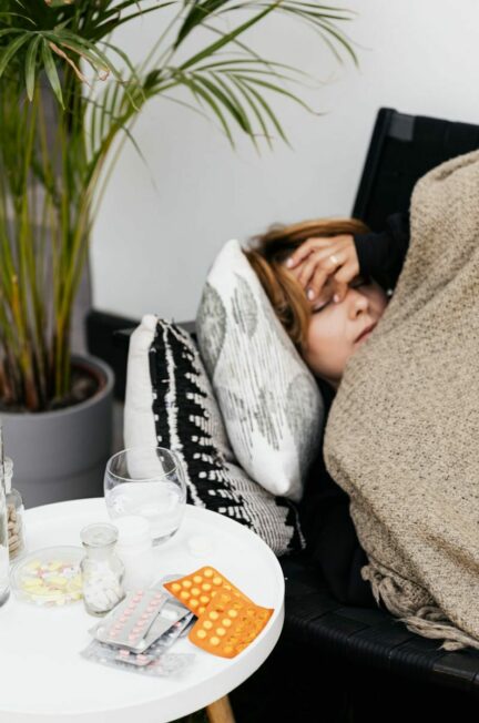 Les comorbidités / Woman lying on a couch covered with a blanket, feeling unwell, with pills on a table nearby.