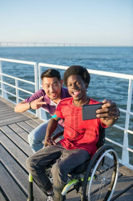 5 dimensions du retablissement - Two friends, one in a wheelchair, taking a selfie on a sunny boardwalk by the water.