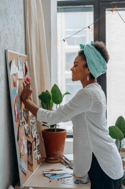 projets maladie chronique / Side view of a smiling woman organizing a cork board with photos in a plant-decorated room.