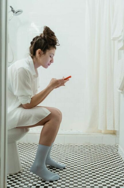 les troubles urinaires - Woman in a bathroom using a smartphone while sitting on the toilet.